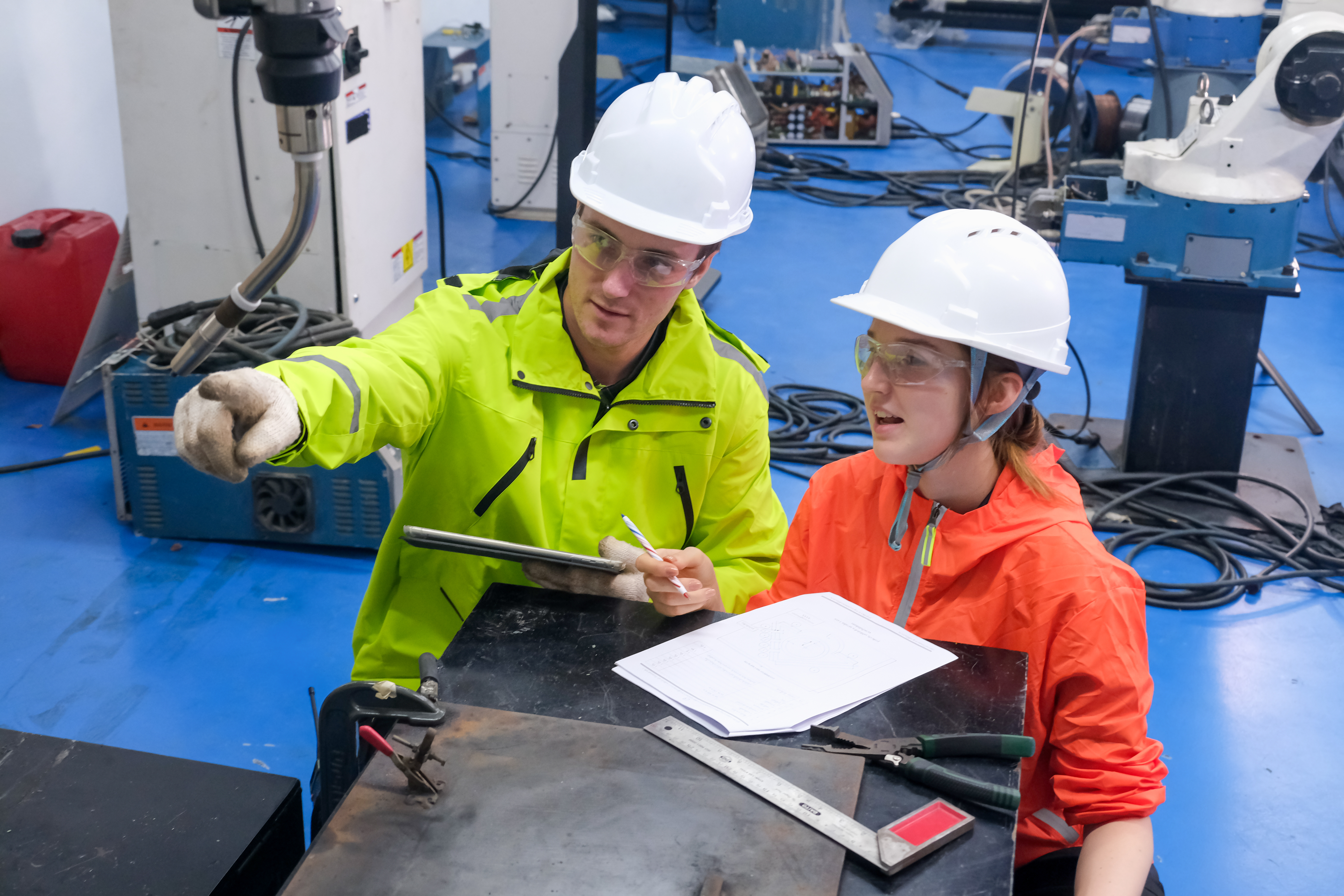 A teacher and student in hardhats and safety goggles discuss machines on a factory floor.